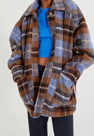 Personne portant une veste surdimensionnée à carreaux marron, bleu et gris avec des boutons, par-dessus un haut court bleu vif et un pantalon foncé à fines rayures.