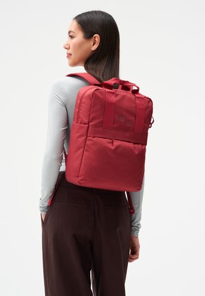 Frau mit langen dunklen Haaren, die einen roten Rucksack, ein hellgraues langärmliges Oberteil und dunkelbraune Hosen trägt, stehend vor einem weißen Hintergrund.