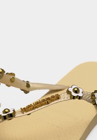 Μπεζ σαγιονάρα με υφασμένους ιμάντες διακοσμημένους με χρυσά και λευκά καρφιά σε σχήμα λουλουδιού και χρυσό λογότυπο της μάρκας.