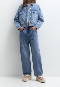 Džínová bunda se dvěma kapsami na hrudi a knoflíky, spárovaná s džíny s vysokým pasem a širokými nohavicemi v světlemodré barvě. Celý vzhled doplňují platformové sandály.