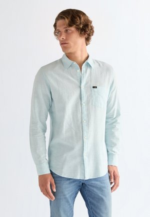 Młody mężczyzna z kręconymi włosami, ubrany w jasnoniebieską koszulę zapinaną na guziki i niebieskie dżinsy, patrzący w bok na tle białego tła.