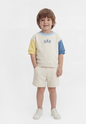 Ung dreng står smilende, iført cremefarvede shorts og en cremefarvet sweatshirt med gule og blå ærmer samt hvide sneakers.