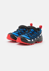 Zapatillas de trail con parte superior de malla azul y negra, suela de goma roja y tiras de Velcro negras. Presentan un diseño texturizado y puntera reforzada.