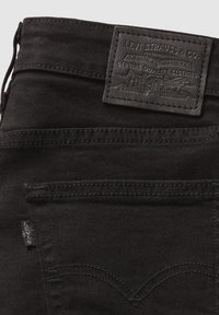 Jeans de mezclilla negra con un parche de cuero texturizado que presenta una marca en relieve. Costuras en contraste visibles a lo largo de los bolsillos y costuras.