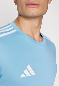 Light blue athletic shirt with a crew neckline; features three white stripes on the shoulder and a white logo on the chest.