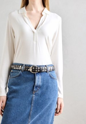 Kobieta ubrana w biały top z długim rękawem i dekoltem w serek, włożony w niebieską spódnicę dżinsową, uzupełniona czarnym paskiem z ćwiekami i srebrną klamrą.