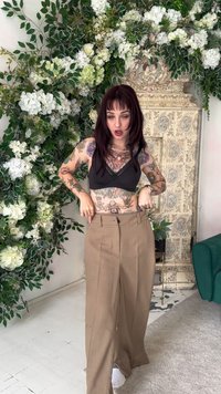 Calças castanhas de cintura alta com um corte solto, apresentando bolsos laterais e uma textura lisa, combinadas com um bralette preto contra um fundo floral.