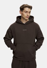 Mørkebrun hettegenser laget av mykt stoff, med kengurulomme, ribbede mansjetter og en liten hvit logo "CASPARA" på brystet.