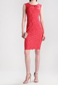 Robe en dentelle rouge avec une silhouette ajustée, des manches courtes et un motif floral. Le mannequin tient une petite pochette et porte des talons à brides.