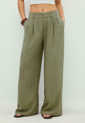 Kobieta nosząca oliwkowo zielone, szerokie spodnie z wysokim stanem, z fałdami, kieszeniami i czarnymi, płaskimi sandałami, stojąca przy jasnej ścianie.