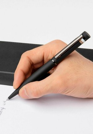 Zwarte pen met een matte afwerking, zilveren accenten, vastgehouden in een hand, schrijft op wit papier, naast een zwart leren notitieboek.