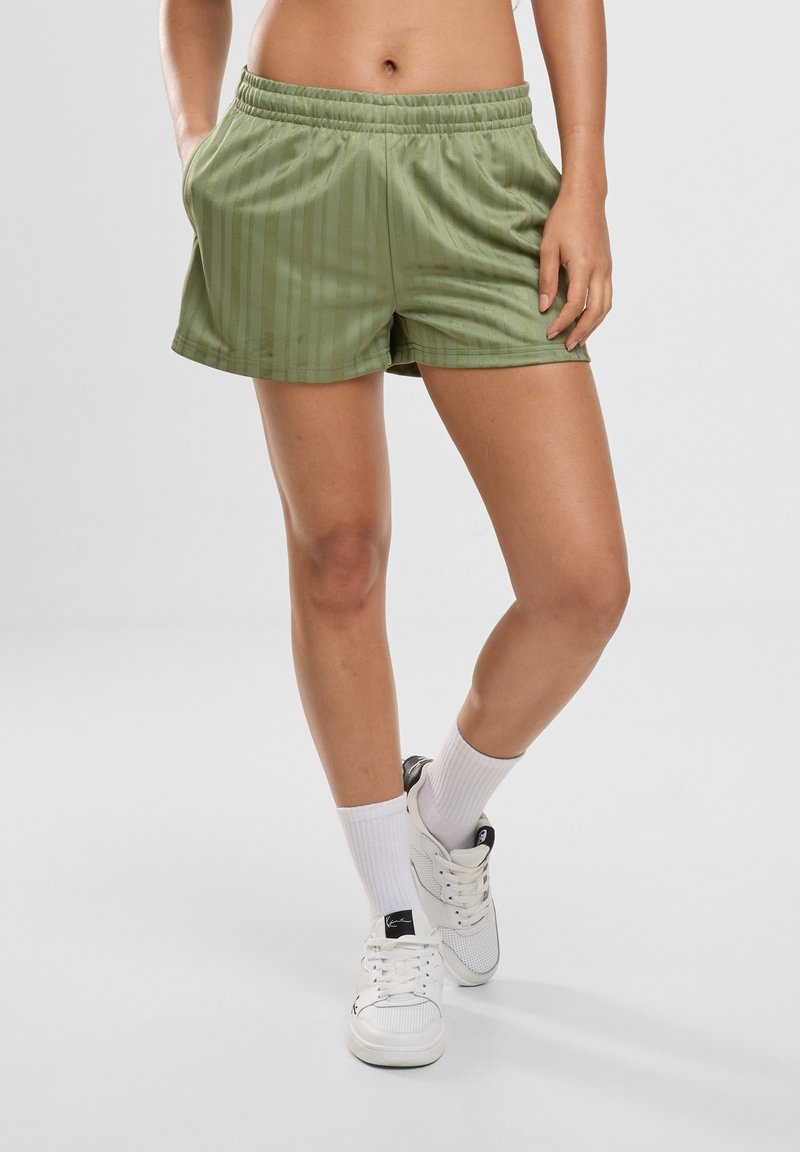 Personne portant un short de sport vert clair avec des poches, des chaussettes blanches montantes et des baskets blanches, debout, une jambe croisée sur l'autre.