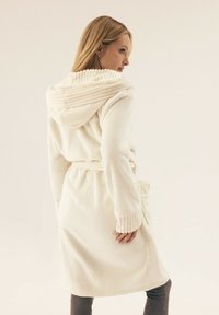 Biała szlafrok z polaru z żebrowanym kapturem, długimi rękawami i paskiem w talii. Posiada boczną kieszeń oraz miękkie, teksturowane wykończenie.