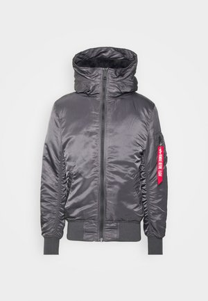 Svart, zip-up bomberjacka med hög krage, ribbade ärmslut och fåll; har en synlig logotypatch och en fäst röd etikett.