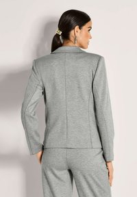 Vrouw met donker haar in een paardenstaart, die zilveren oorbellen draagt en een lichtgrijze, getailleerde blazer met bijpassende broek, met de rug naar de camera tegen een effen achtergrond.