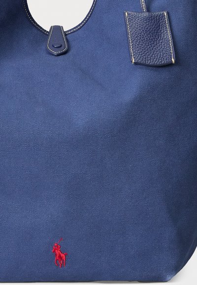 Marinblå canvas tote-väska med läderdetaljer. Har en röd broderad logotyp av en häst med en ryttare. Rundad utskärning på handtaget och strukturerad tyg.