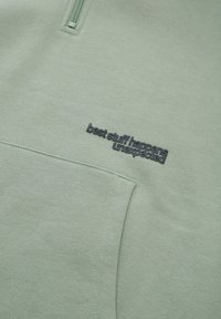 Felpa verde menta con collare a mezza zip, caratterizzata da un logo nero texturizzato che recita "le migliori cose accadono inaspettatamente" sul davanti.
