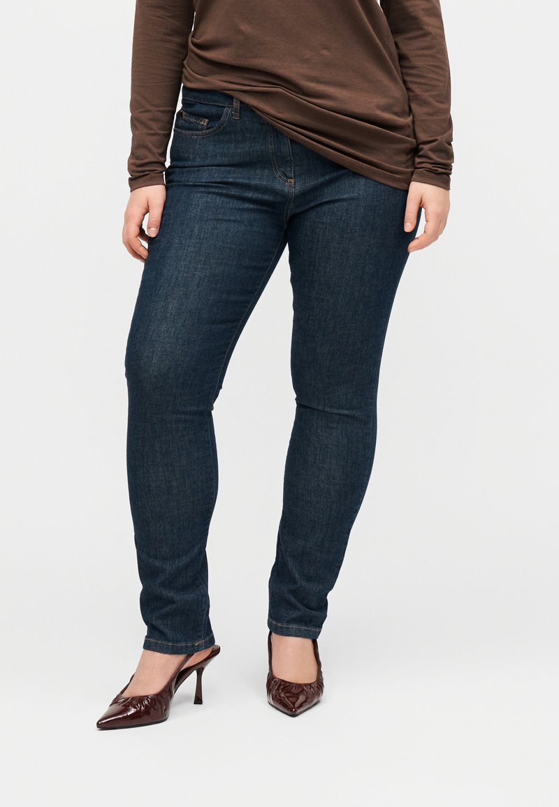 Άτομο που φοράει σκούρο μπλε στενά τζιν παντελόνια, συνδυασμένα με καφέ μακρυμάνικη μπλούζα και σκούρα καφέ ψηλοτάκουνα παπούτσια με μυτερή μύτη.