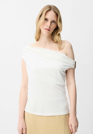 Femme aux cheveux blonds portant un haut blanc asymétrique avec un détail boucle sur le côté droit et une jupe beige, sur fond blanc.