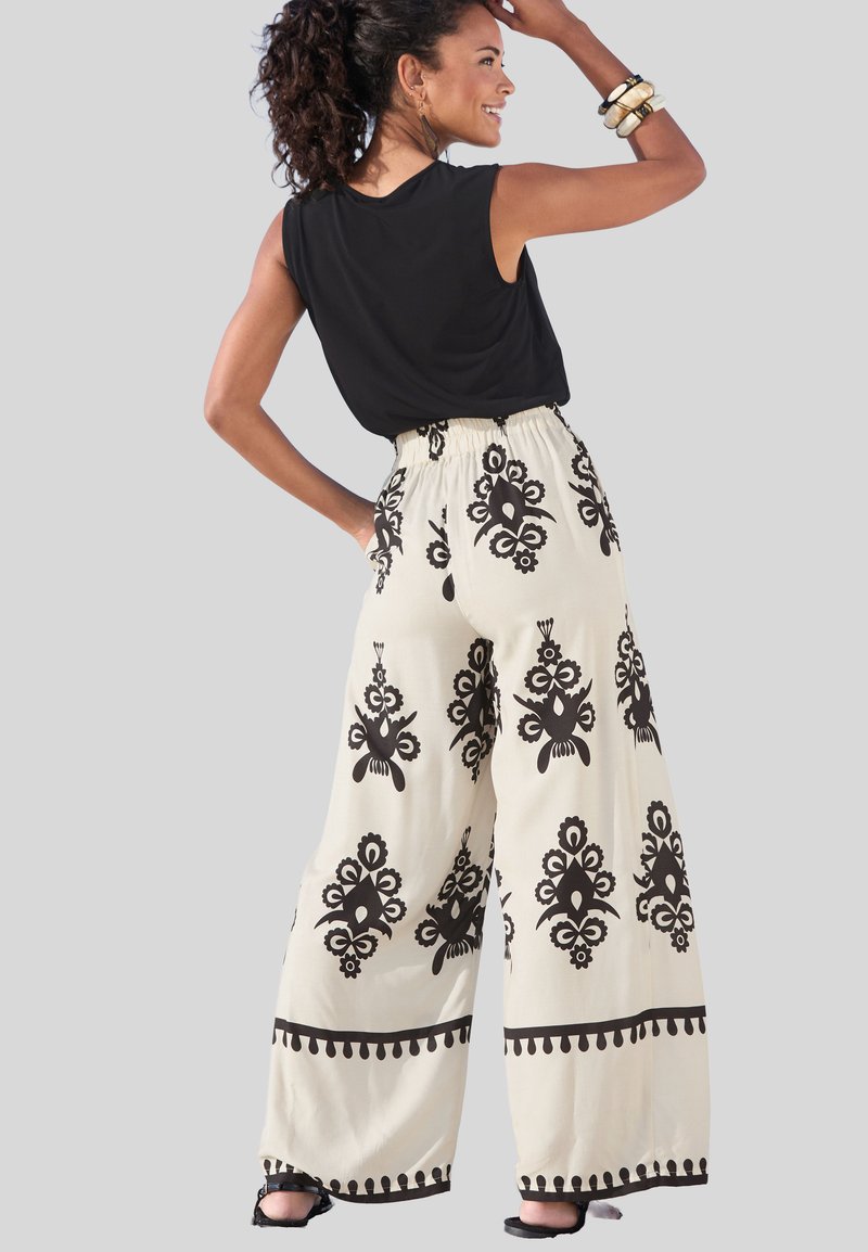 Wide-leg pants feature a cream background with bold black floral patterns, an elastic waistband, and a decorative trim at the hem.