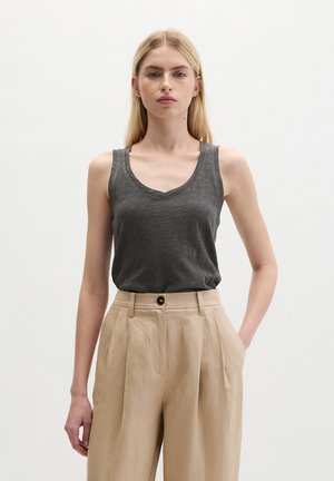 Blonde woman wearing sleeveless dark gray V-neck top tucked into high-waisted beige pleated trousers, standing against plain background.
