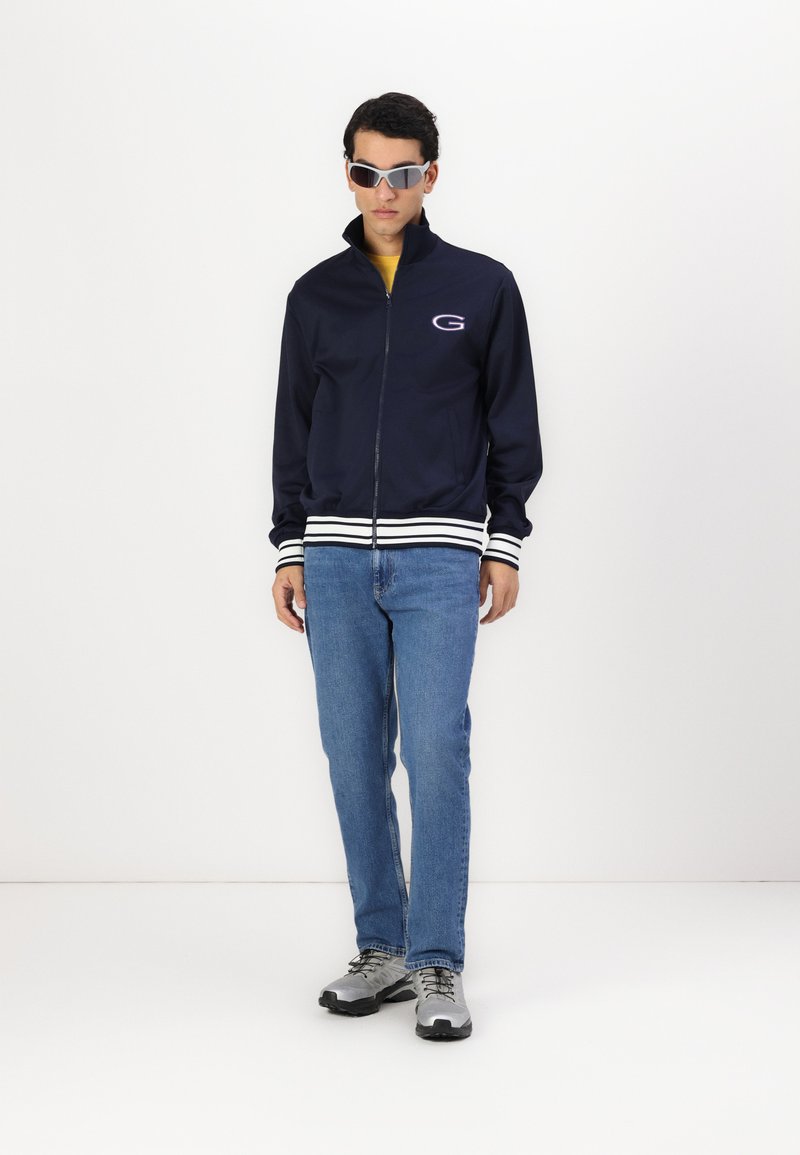 Chaqueta de chándal con cremallera en color navy y rayas blancas, logo en el pecho. Combinada con jeans azules y zapatillas deportivas grises. Modelo usando gafas de sol.