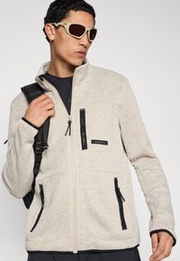 Μπεζ υφασμάτινο σακάκι με φερμουάρ μπροστά, μαύρες λεπτομέρειες και πολλαπλές τσέπες με φερμουάρ. Φορεμένο πάνω από μαύρο πουκάμισο, με σακίδιο στον έναν ώμο.