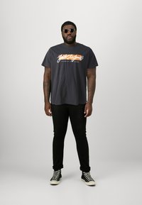 Black graphic t-shirt with "Jack & Jones" logo in white and orange. Pair of black jeans and black high-top sneakers. Circular sunglasses.
