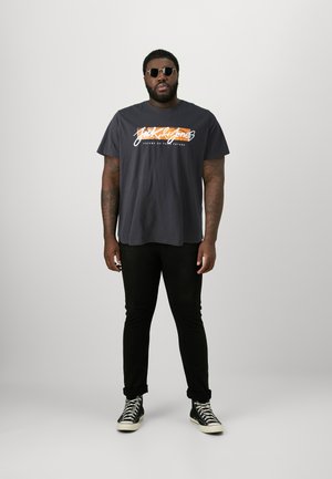 Camiseta gráfica negra con el logo "Jack & Jones" en blanco y naranja. Un par de jeans negros y zapatillas de deporte negras de caña alta. Gafas de sol circulares.