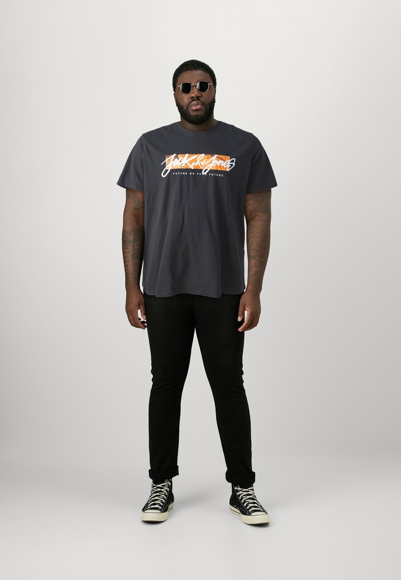 Black graphic t-shirt with "Jack & Jones" logo in white and orange. Pair of black jeans and black high-top sneakers. Circular sunglasses.