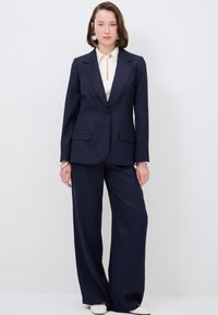 Blazer ajusté bleu marine avec un bouton, revers en dents de scie, et deux poches avant, associé à un pantalon large et une chemise blanche.