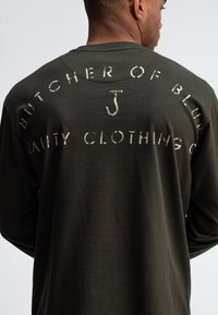 Langärmliges olivgrünes Hemd mit weißem Drucktext auf der Rückseite, der "BUTCHER OF BLUE" und "VANITY CLOTHING CO" in einer lässigen Schriftart sagt.