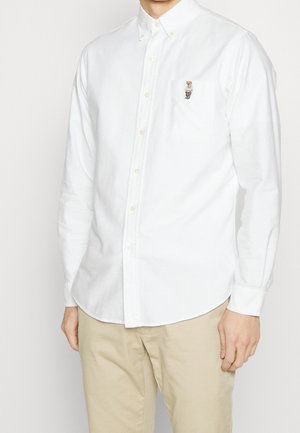 Mand iført en hvid skjorte med knapper og en lille broderet bamse på brystlommen samt beige bukser.