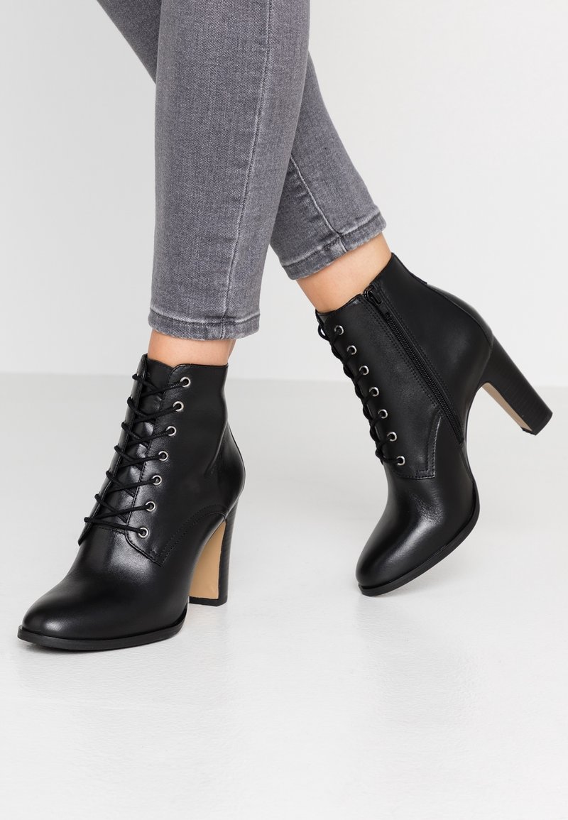 Bottines noires en cuir à lacets avec fermeture éclair sur le côté et talons blocs hauts, portées avec un jean gris ajusté sur un sol blanc.