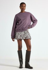 Donna in posa con un ampio maglione viola, una mini gonna floreale, orecchini a cerchio dorati e stivali neri in pelle al ginocchio su uno sfondo bianco.