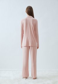 Rosa långärmad topp och matchande byxor i mjukt tyg. Toppen har krage, medan byxorna har vid fåll. Texturerad bakgrund.