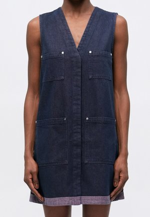 Robe sans manches en denim foncé avec un col en V et quatre poches devant, portée par une personne avec les bras et les mains visibles.