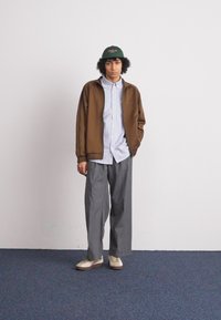 Veste marron sur une chemise rayée bleue et blanche, pantalon large gris, et chaussures crème. Accessoire : casquette verte. Fond uni.