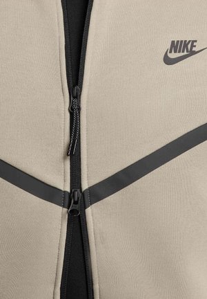 Beżowa kurtka zapinana na zamek Nike z czarnym suwakiem i akcentami. Posiada ukośne czarne paski. Gładka tekstura, sportowy fason.