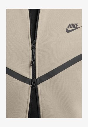 Beżowa kurtka zapinana na zamek Nike z czarnym suwakiem i akcentami. Posiada ukośne czarne paski. Gładka tekstura, sportowy fason.