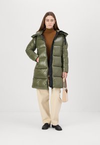 Olijfgroene gewatteerde jas met een glanzende afwerking, zwarte accenten en een capuchon. Het model draagt een bruine gebreide trui en crèmekleurige wijde pantalons, en houdt een handtas vast.