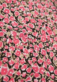 Blommönstrad tyg med rosa och beige blommor på en svart bakgrund; har knappar och en fickdetalj.