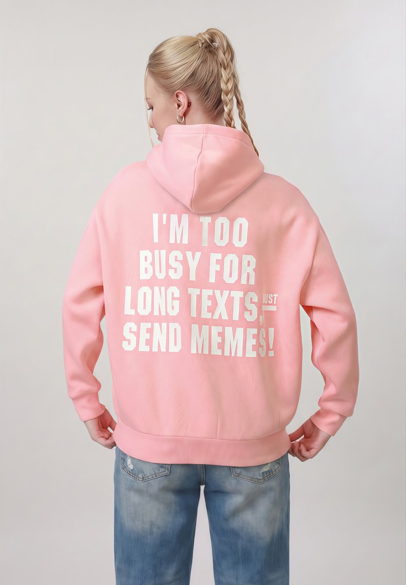 Personne aux cheveux blonds tressés portant un sweat à capuche rose avec un texte blanc : "JE SUIS TROP OCCUPÉ(E) POUR LES LONGS TEXTES, ENVOYEZ JUSTE DES MÈMES !"