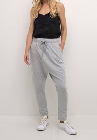 Vrouw in losse grijze broek met trekkoord, zwarte mouwloze top met kantdetails en witte sneakers, staande tegen een effen achtergrond.