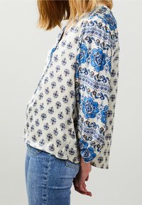 Blusa bianca con motivi floreali blu, con scollo a V e maniche lunghe, realizzata in un tessuto leggero. Abbinata a jeans in denim blu.