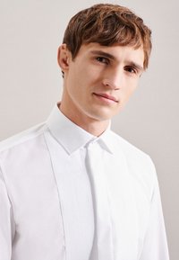 Camicia bianca con design a pieghe sul davanti, colletto classico, texture liscia e silhouette aderente.