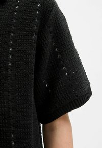 Top noir en tricot avec des manches courtes et amples. Le tissu texturé présente un motif de petits trous et un ourlet côtelé au niveau des manches.