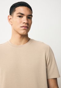 T-shirt beige en coton avec un col rond, des manches courtes et une texture lisse, présentant des détails de couture minimalistes.