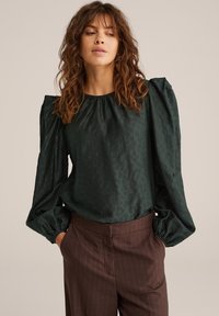 Blouse vert foncé avec des manches bouffantes, en tissu texturé et un col rond, associée à un pantalon marron à rayures.