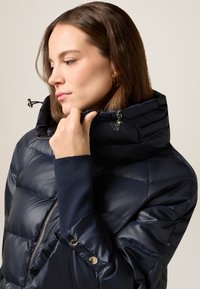 Marineblå puffjakke med h�øj krave, skinnende tekstur, lynlåslukning og trykknapsdetaljer. Har tætsiddende ærmer med et ensfarvet underlag.